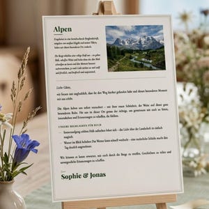 May include: A printed card on a small wooden easel, featuring the word "Alpen" at the top. The card includes text and a scenic image of mountains and a river. The card is decorated with a small vase of flowers.