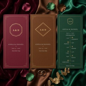 May include: Three wedding invitation cards in burgundy, brown, and emerald green. Each card features gold geometric designs, the initials "A & D", and the names "Anna & Daniel" with the date "14 June 2026". The green card includes a wedding day schedule.