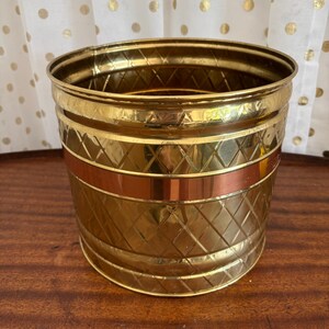 May include: A cylindrical brass planter featuring a polished finish with a diamond-patterned texture across the exterior surface. A smooth, horizontal copper-colored band encircles the middle section of the vessel, providing a contrast to the gold-toned metal.
