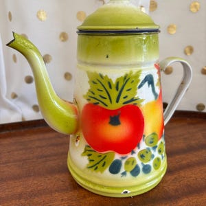 May include: An enamel coffee pot with a cream-colored body and a light green lid, base, and spout. The exterior features a colorful painted design of a large red apple, green leaves, and small clusters of green and dark blue grapes.