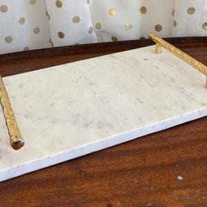 May include: A rectangular white marble tray featuring a smooth, polished surface with natural grey veining. The tray is equipped with two gold-toned metal handles that have a textured, branch-like appearance on each of the shorter sides.