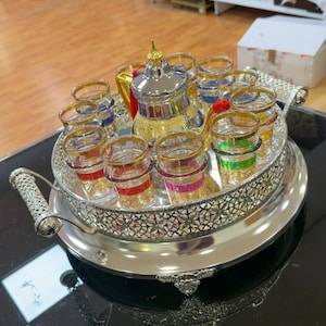 Puede incluir: Un juego de té plateado con una tetera y varios vasos pequeños, cada uno con una banda de color diferente. El juego está sobre una bandeja plateada decorativa con asas. La tetera tiene una tapa dorada.