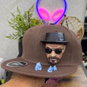 Può includere: Un cappellino da baseball marrone con una figura tridimensionale stampata di un uomo con cappello nero e occhiali da sole fissata sul pannello frontale. Tre piccoli accenti azzurri a forma di cristallo geometrico sono sulla visiera piatta.