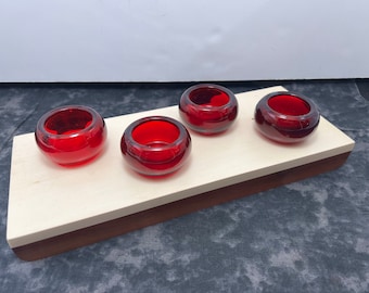 May include: A wooden tray with four small, round, red glass candle holders. The tray has a light-colored top and a dark brown base. The candle holders are evenly spaced across the tray.