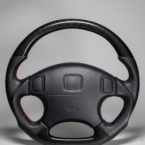 Könnte beinhalten: Schwarzes Lenkrad mit Kohlefaserakzenten und roten Nähten. Das Lenkrad hat einen perforierten Ledergriff und ein Honda-Emblem in der Mitte. Enthält zwei Bedientasten und ein SRS-Airbag-Etikett.