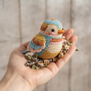 May include: A small, round, crocheted bird figurine featuring a color palette of light blue, soft pink, mustard yellow, and cream. The bird has black bead eyes, a small orange beak, and is perched on a textured brown branch with tiny flowers.