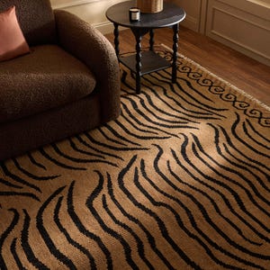 May include: A rectangular area rug featuring a tan base colour with a repeating black tiger stripe pattern and a decorative border design along the edge. The rug has a textured surface and includes short fringe detailing along the short side edge.