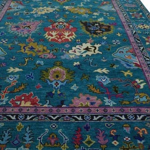 May include: A teal-colored area rug with an intricate floral pattern. The rug features a central field of teal with colorful floral motifs in pink, yellow, red, and blue. A border of pink and black surrounds the central design. A woven basket is in the upper left corner.