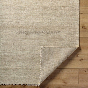 May include: Beige rectangular rug with a textured surface. The corner of the rug is folded up, revealing the underside. The rug is on a wooden floor. The text "VintageRugHouseArt" is visible on the rug.