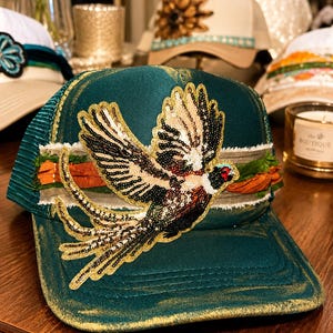Puede incluir: Una gorra tipo trucker de color verde azulado oscuro con una gran aplicación de faisán con lentejuelas en el panel frontal. La gorra incluye una banda horizontal de tela deshilachada en tonos naranja y gris, con detalles dorados en la visera.