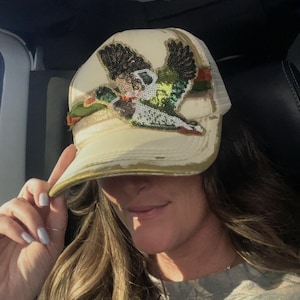 Puede incluir: Una gorra tipo camionero de color crema con un parche de lentejuelas de un pato real en vuelo. La gorra tiene una visera desgastada, una parte trasera de malla y una banda dorada en el frente. El diseño del pato incluye lentejuelas verdes, marrones y blancas.
