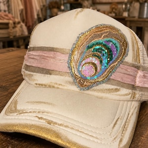 May include: A distressed cream-colored baseball cap featuring a horizontal pink fabric band and a large, central embroidered patch shaped like an oyster shell. The patch is decorated with iridescent sequins in shades of blue, green, and pink.