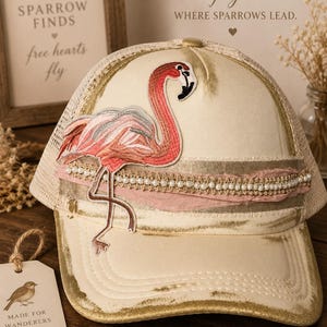 Puede incluir: Una gorra de béisbol color crema con acabado dorado desgastado, que presenta un parche bordado de un flamenco rosa. La gorra está decorada con una banda horizontal de perlas blancas y ribetes de tela deshilachada, e incluye un panel trasero de malla.