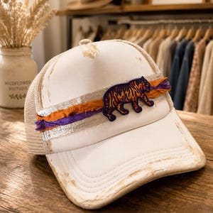 May include: A distressed white trucker cap featuring a mesh back and a structured front panel. The front is decorated with a horizontal silver metallic band, layered with frayed orange and purple fabric strips, and a tiger patch in purple and orange.