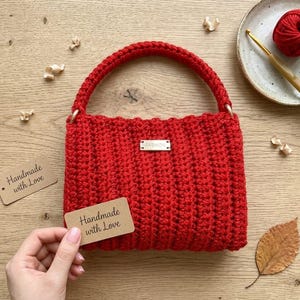 May include: A handmade red crochet handbag with a structured design and a top handle. The bag features a small metal tag that reads "FASHION". A tag that reads "Handmade with Love" is held near the bag. The bag is approximately 8 inches wide.