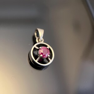 Könnte beinhalten: Ein silberfarbener Anhänger mit einem kreisförmigen Rahmen und einer kreuzförmigen Fassung, die einen einzelnen, runden, facettierten, tief rosa-roten Edelstein in der Mitte hält. Der Anhänger verfügt über eine kleine Öse an der Oberseite.
