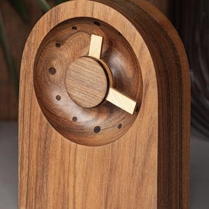 Puede incluir: Un reloj de madera hecho a mano con un diseño único. El reloj presenta una esfera circular con un dial central y tres manecillas. El reloj está hecho de madera de tonos cálidos, con un patrón de vetas natural. El reloj mide unos 15 cm de alto.