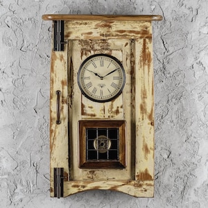 Puede incluir: Un reloj de pared crema desgastado con una estética vintage. El reloj presenta una esfera redonda con números romanos y manecillas negras. Debajo de la esfera, una pequeña ventana cuadrada con un diseño metálico decorativo. El reloj está montado en un marco de madera.