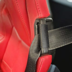 May include: A bright red car seat with a textured surface and visible stitching, featuring a black plastic seatbelt guide attached to the side. A black woven seatbelt strap passes through the guide, extending horizontally across the frame.