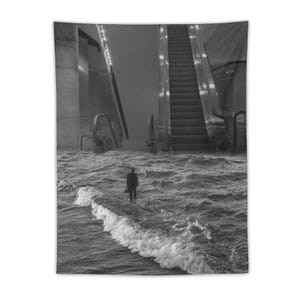 May include: A black and white wall tapestry featuring a surreal scene where a person in a suit stands on the surface of choppy ocean waves. In the background, a large metal escalator rises from the water towards a dark, indistinct sky.