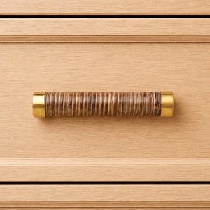 May include: A light-coloured wooden drawer with a cylindrical handle. The handle is wrapped in a brown, textured material and has gold-coloured metal caps on each end. The drawer is part of a larger piece of furniture.