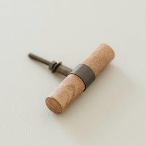 Può includere: Un pomello per mobili cilindrico in legno con una finitura naturale a venature color marrone chiaro. Il pomello è accentuato da una fascia metallica scura attorno al centro e include un bullone filettato sul retro per il fissaggio.