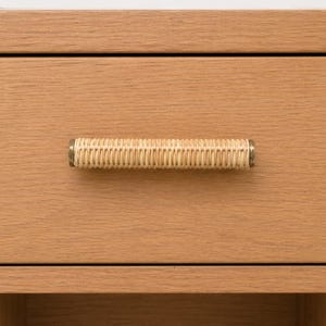 May include: Close-up of a wooden drawer with a woven rattan handle. The drawer is a light brown color and has a simple, clean design. The handle is cylindrical with brass-colored ends, adding a touch of elegance to the piece.