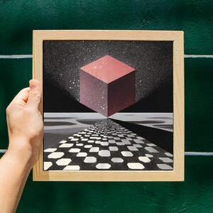 Pode incluir: Uma obra de arte emoldurada com um design geom&eacute;trico. Um cubo vermelho flutua sobre um caminho quadriculado em preto e branco, contra um c&eacute;u estrelado. A obra de arte &eacute; segurada por uma m&atilde;o, com uma moldura de madeira e um fundo verde.