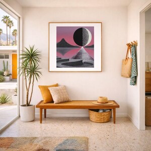 K&ouml;nnte beinhalten: Ein gerahmter Kunstdruck zeigt eine geometrische Landschaft mit einem schwarz-wei&szlig;en Mond, Bergen und Wasser vor einem rosa-violetten Himmel. Das Kunstwerk h&auml;ngt &uuml;ber einer Holzbank mit Kissen, einem Strohhut und einem geflochtenen Korb.
