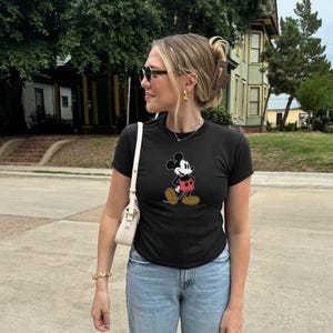 May include: Black t-shirt featuring a classic Mickey Mouse graphic. The cartoon character is depicted in his iconic outfit with red shorts, white gloves, and brown shoes. The shirt is paired with light blue jeans.