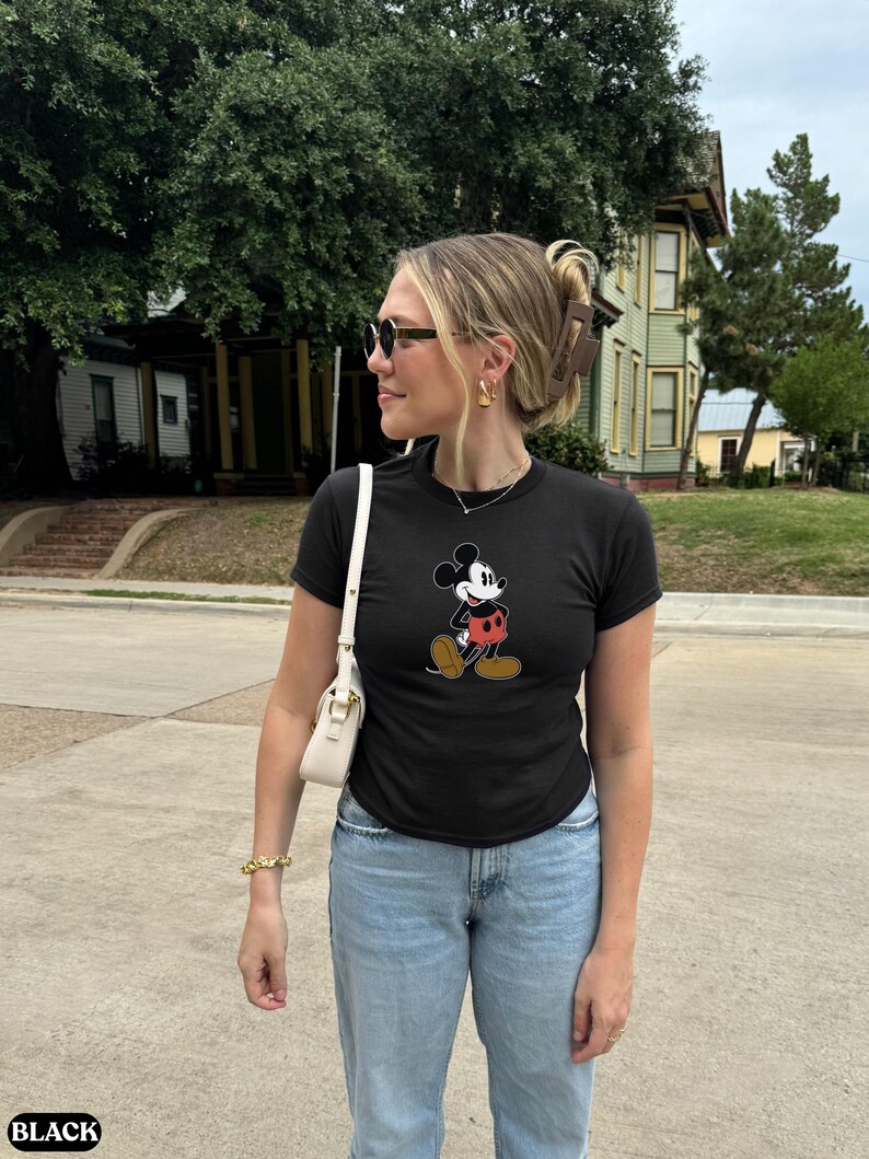 May include: Black t-shirt featuring a classic Mickey Mouse graphic. The cartoon character is depicted in his iconic outfit with red shorts, white gloves, and brown shoes. The shirt is paired with light blue jeans.