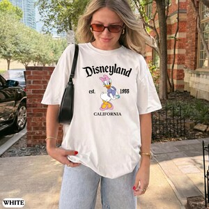 May include: White t-shirt with the Disneyland logo and a Daisy Duck graphic. The graphic includes the text "est. 1955 CALIFORNIA". The shirt is a casual style.