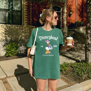 May include: Light green t-shirt with the Disneyland logo, Daisy Duck graphic, and "California" text. The shirt is a relaxed fit and worn with a small white purse. The image is taken outdoors on a sunny day.