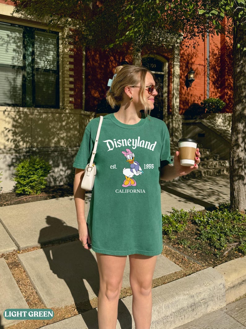 May include: Light green t-shirt with the Disneyland logo, Daisy Duck graphic, and "California" text. The shirt is a relaxed fit and worn with a small white purse. The image is taken outdoors on a sunny day.