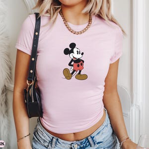 May include: Light pink t-shirt featuring a classic Mickey Mouse graphic. The cartoon character is depicted in black, red, and brown, walking with a cheerful expression. The shirt is short-sleeved and appears to be made of a soft material.