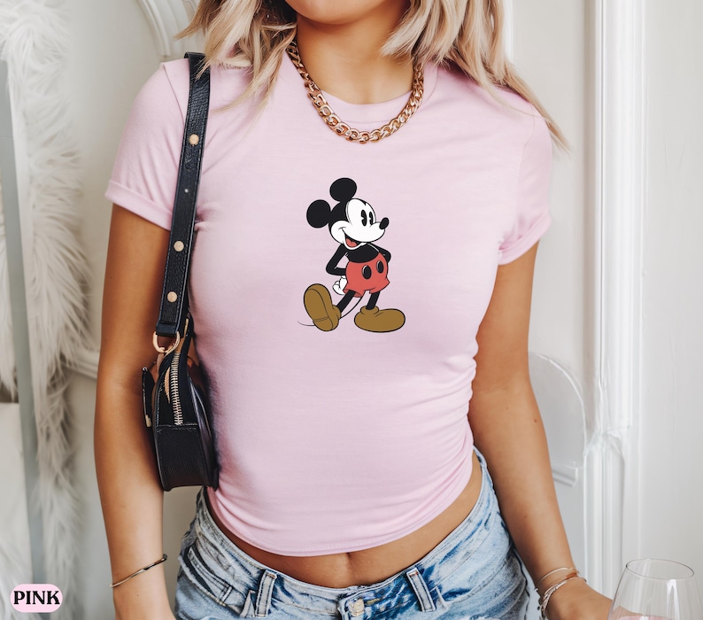 May include: Light pink t-shirt featuring a classic Mickey Mouse graphic. The cartoon character is depicted in black, red, and brown, walking with a cheerful expression. The shirt is short-sleeved and appears to be made of a soft material.