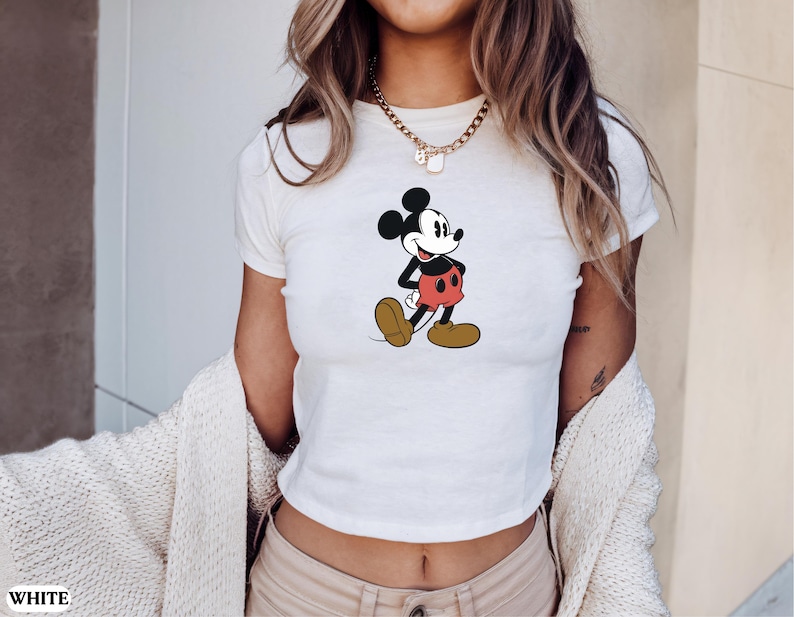 May include: White crop top featuring a classic Mickey Mouse graphic. The cartoon character is depicted in his signature pose, wearing red shorts, yellow shoes, and black gloves. The shirt is a casual, everyday wear item.