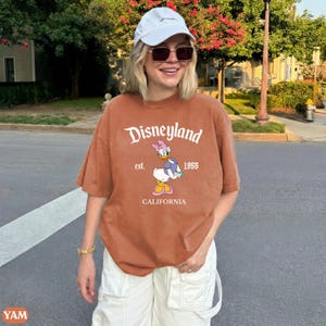 May include: A rust-colored t-shirt with the Disneyland logo and Daisy Duck graphic. The shirt includes the text "est. 1955 CALIFORNIA". The shirt is paired with white shorts and a white baseball cap.