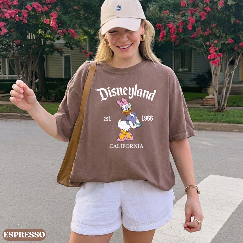 May include: Espresso-colored t-shirt with the Disneyland logo, Daisy Duck graphic, and "est. 1955 CALIFORNIA" text. The shirt is paired with white shorts and a beige baseball cap.