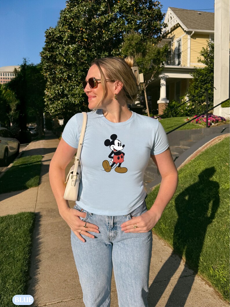 May include: Light blue t-shirt featuring a classic Mickey Mouse graphic. The shirt has short sleeves and a crew neck. The person is wearing light wash blue jeans and sunglasses. A white purse is hanging from the shoulder.