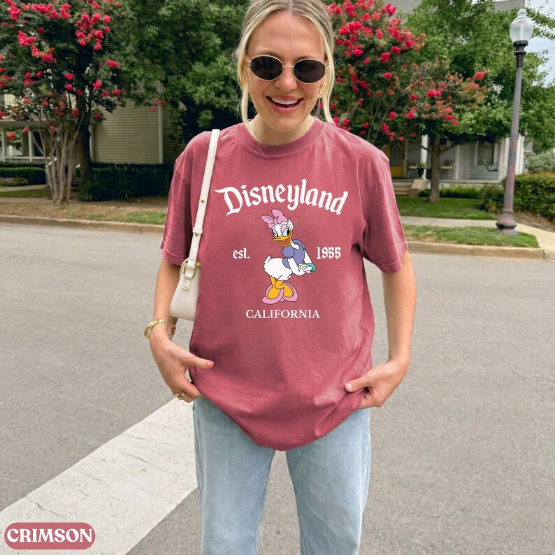May include: A maroon t-shirt with the Disneyland logo and Daisy Duck graphic. The shirt includes the text "est. 1955 CALIFORNIA". The shirt is a short-sleeved crew neck.