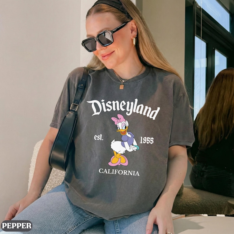 May include: Gray t-shirt featuring the Disneyland logo with Daisy Duck. The shirt includes the text "est. 1955" and "CALIFORNIA" below the graphic. The shirt is a casual style.