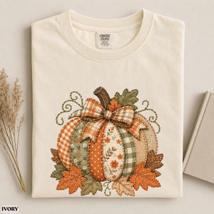 May include: An ivory t-shirt featuring a central graphic of a pumpkin composed of various fabric-like patterns including orange gingham, green plaid, floral prints, and polka dots, topped with a plaid bow and surrounded by autumn leaves.