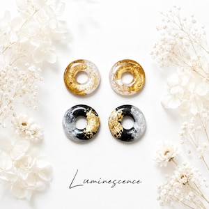 May include: Four circular resin ornaments arranged in a square formation. The top two are clear with gold leaf flakes, while the bottom two are black and grey with gold leaf accents. The word Luminescence is printed in black script below them.