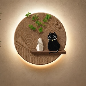 Könnte beinhalten: Eine runde Wanddekoration mit einem strukturierten, hellbraunen Hintergrund und einem warmen, leuchtenden Licht. Sie zeigt ein kleines Holzregal mit einer weißen Vase, grünen Zweigen und einer schwarzen Katzenfigur. Die Katze hat ein weißes Halsband.