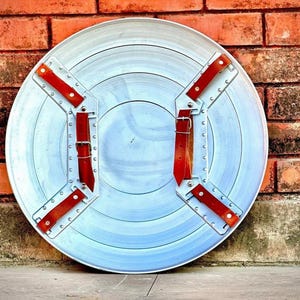 May include: A silver-colored, circular shield with concentric rings and red leather straps. The shield has a metal frame with rivets and is set against a brick wall. The shield's diameter is approximately 24 inches.