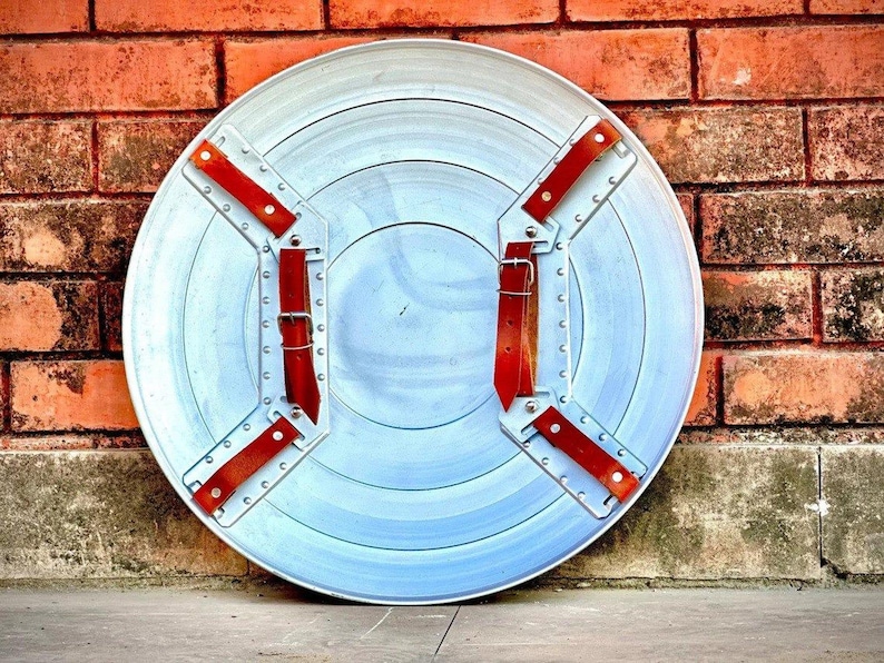 May include: A silver-colored, circular shield with concentric rings and red leather straps. The shield has a metal frame with rivets and is set against a brick wall. The shield's diameter is approximately 24 inches.