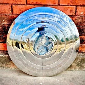 May include: A shiny, silver shield with a five-pointed star in the center. The shield has concentric circular ridges and reflects the surrounding environment. It is leaning against a red brick wall.