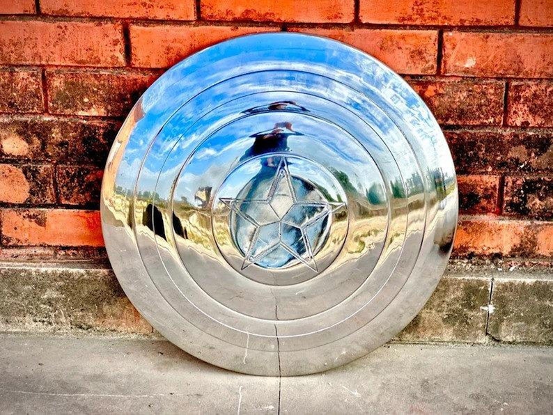 May include: A shiny, silver shield with a five-pointed star in the center. The shield has concentric circular ridges and reflects the surrounding environment. It is leaning against a red brick wall.