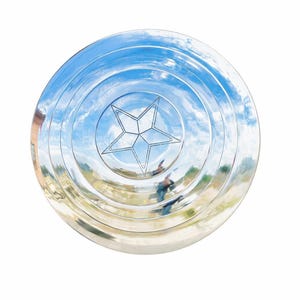 May include: A shiny, silver shield with a five-pointed star in the center. The shield has concentric circles and reflects a blue sky with clouds. A classic superhero accessory.